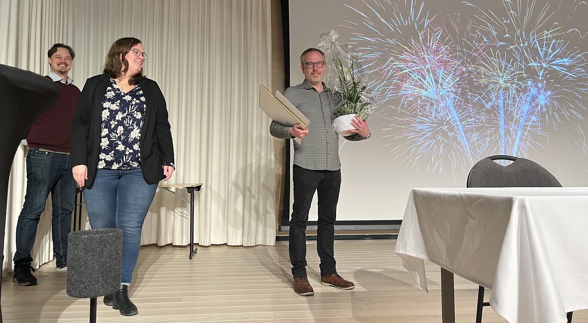 Mats Börjesson står med blomma och diplom framför en bild med fyrverkerier. I vänstra kanten syns politikerna.